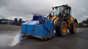 The Street Washing Unit is attached to a yellow-colored wheel loader (model JCB 419), which is driving forward and cleaning the street.