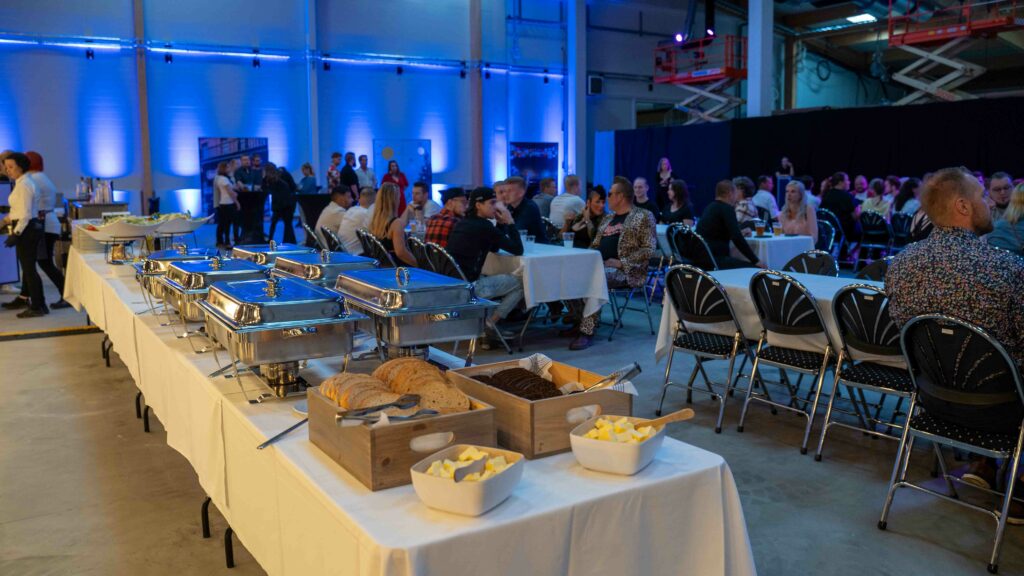 Photo shows buffet table in the foreground, a basket of bread and food in containers. In the background, tables with people seated to celebrate and eat. 