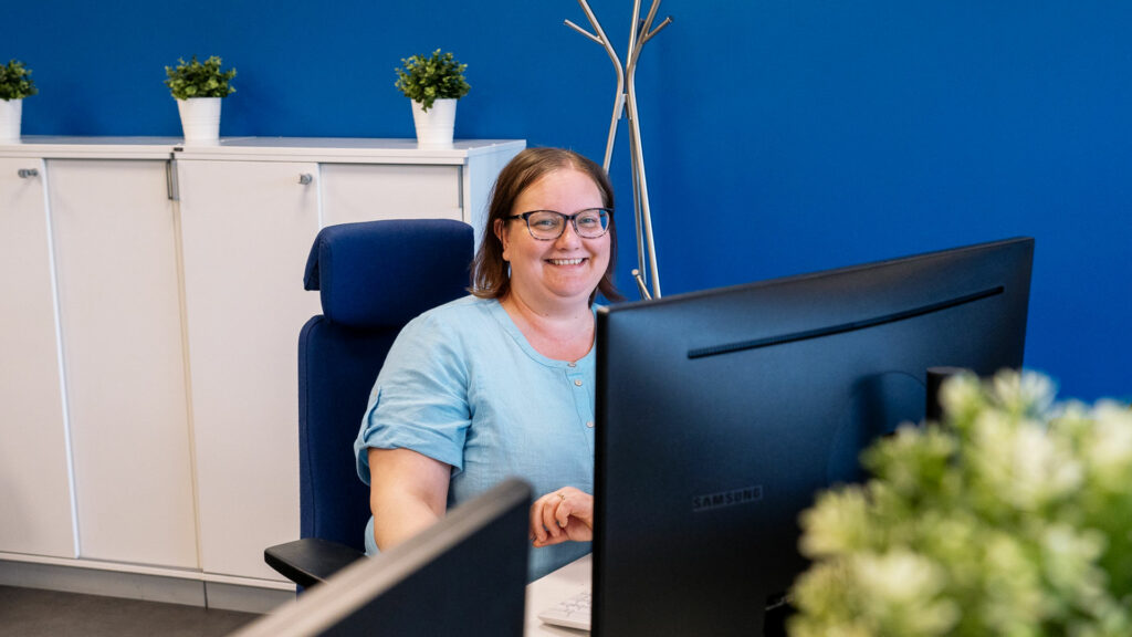 The person sits at her desk and smiles at the camera. In the foreground you can see a plant. In the background are a blue wall and a white cabinet with green plants.